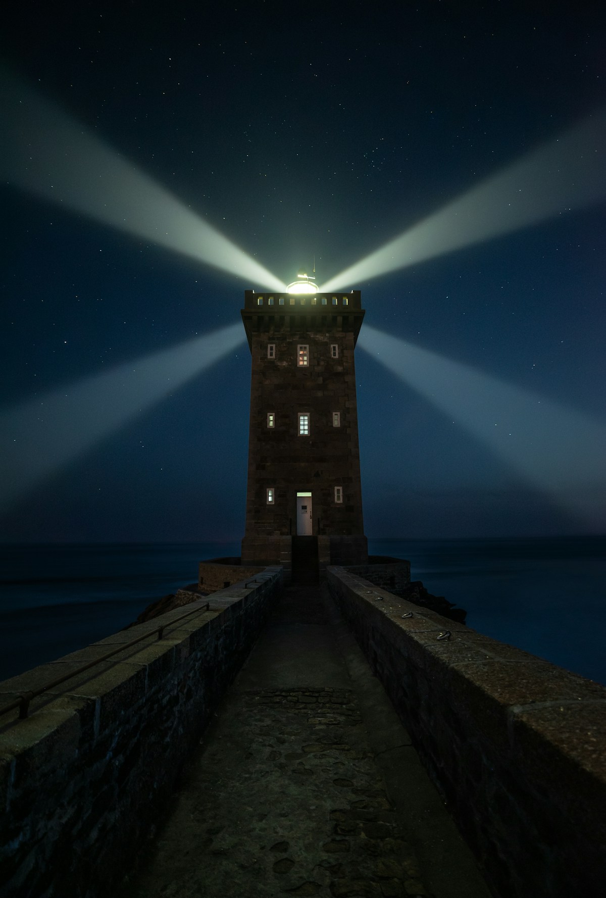 Lighthouse beam sweeping across a starry night sky, stone tower on a narrow walkway over dark ocean